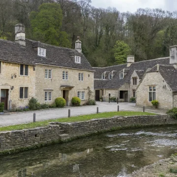 A charming view of a quaint village featuring traditional stone houses with lush greenery, situated beside a serene stream with a small bridge.