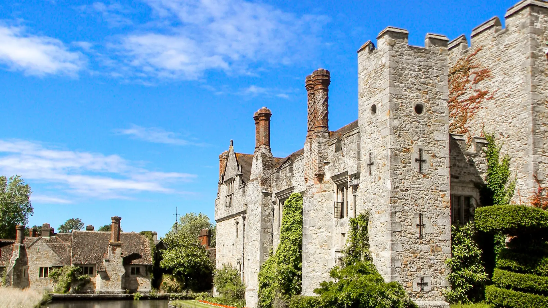 An ancient stone castle with tall chimneys and ivy-covered walls under a clear blue sky, surrounded by lush greenery and a calm moat.