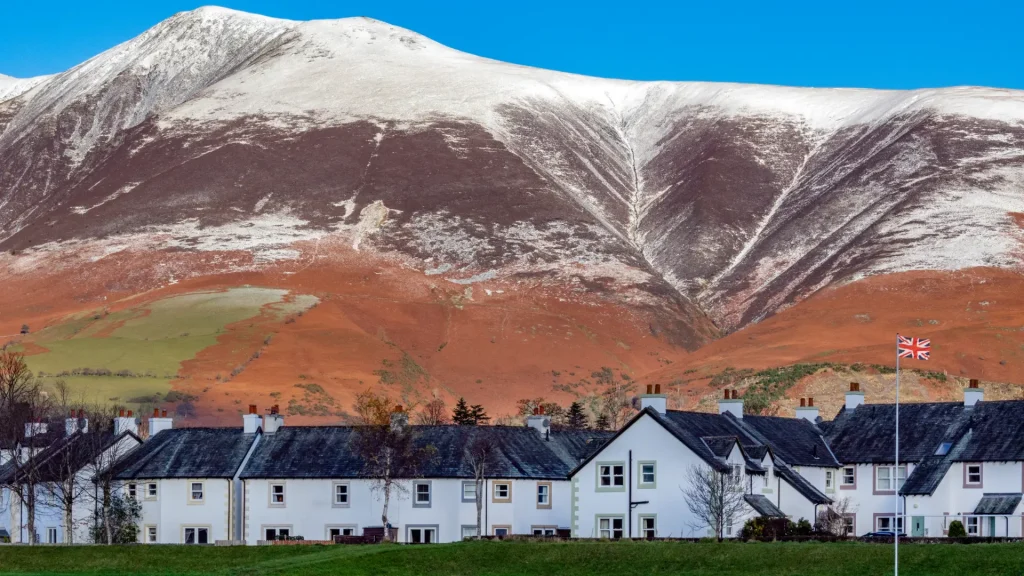 A row of white houses with red roofs in the foreground, a green grassy field in the middle, and snow-capped mountains in the background under a clear blue sky. a british flag flies on the left.