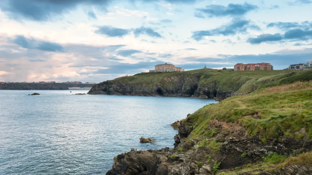 Coastal landscape with rocky cliffs and green grass leading to buildings on a headland under a cloudy sky, overlooking a calm sea.