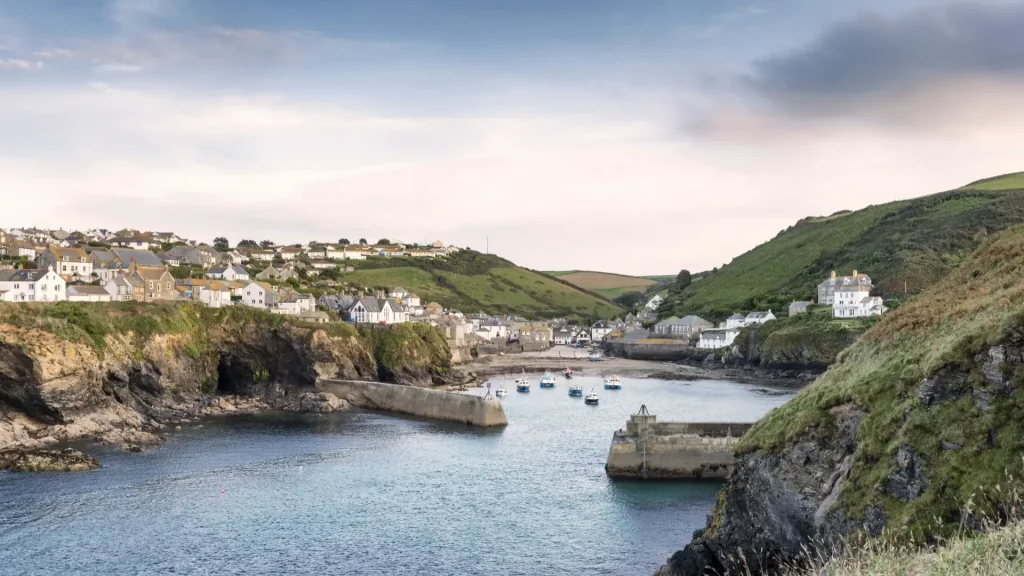 A scenic view of a coastal village with white houses nestled on rolling hills. a harbor with boats is flanked by natural cliffs under a soft sky.