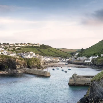 A scenic view of a coastal village with white houses nestled on rolling hills. a harbor with boats is flanked by natural cliffs under a soft sky.