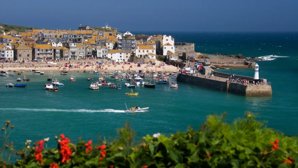A picturesque coastal village with colorful buildings along a beachfront crowded with people. a harbor with boats and a pier extends into vibrant blue sea, framed by greenery and clear skies.