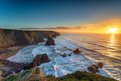Sunset over a rugged coastline with cliffs and rock formations, waves crashing against the shore and reflecting the sunlight, under a clear sky.