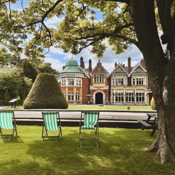 Three green-striped deck chairs face a lavish historic building with intricate brickwork and a green copper dome, set in a well-manicured garden with a large tree foreground.