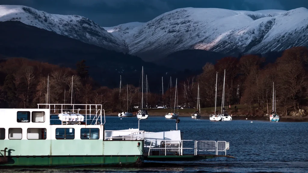A serene lake scene with a ferry in the foreground and sailboats dotted around. snow-capped mountains under a moody sky serve as the backdrop.