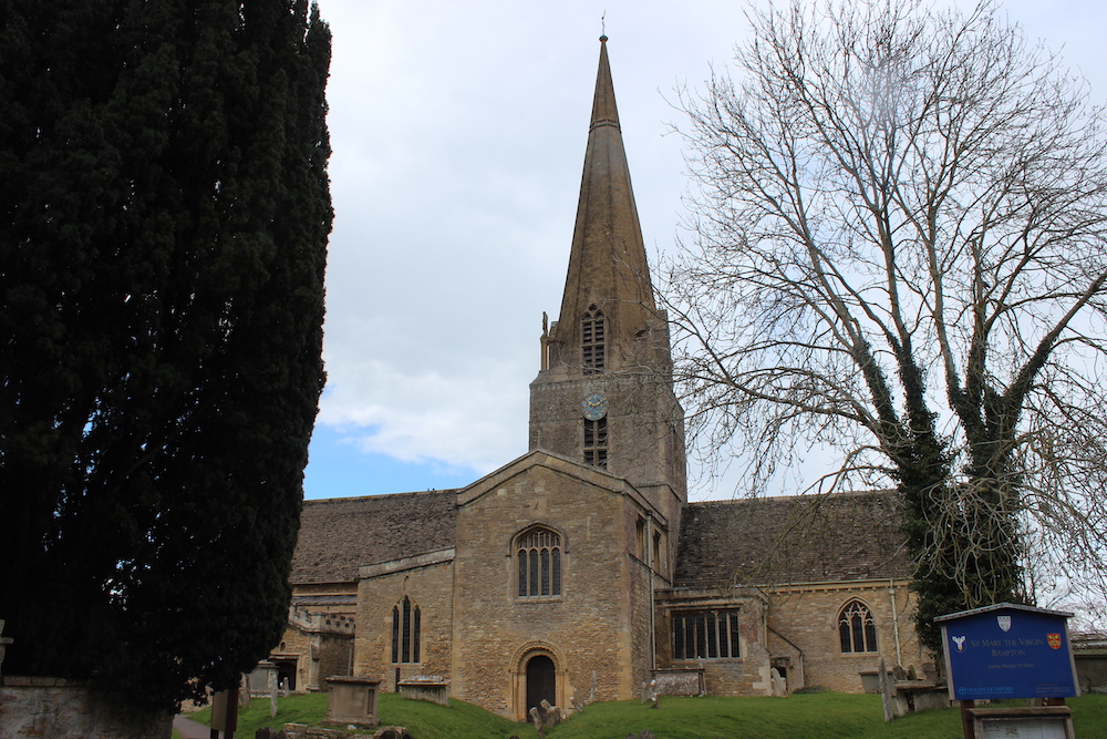 A traditional stone church with a tall spire, featuring a blue clock face, surrounded by trees under a cloudy sky. a signboard is visible near the entrance.