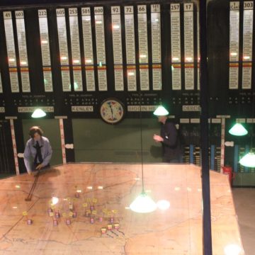 A recreation of a wwii-era war room with a large map table and wall boards displaying flight squadrons under green lamp lights, featuring actors in period costumes.