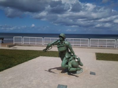 Bronze sculpture of two soldiers in combat gear, one helping the other, on a concrete platform. in the background, there's a sandy beach, ocean, and partly cloudy sky visible within a metal railing.
