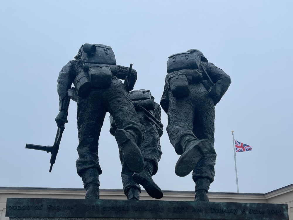 Statue depicting three soldiers in motion, carrying heavy backpacks and equipment under a cloudy sky, with a british flag flying in the background.