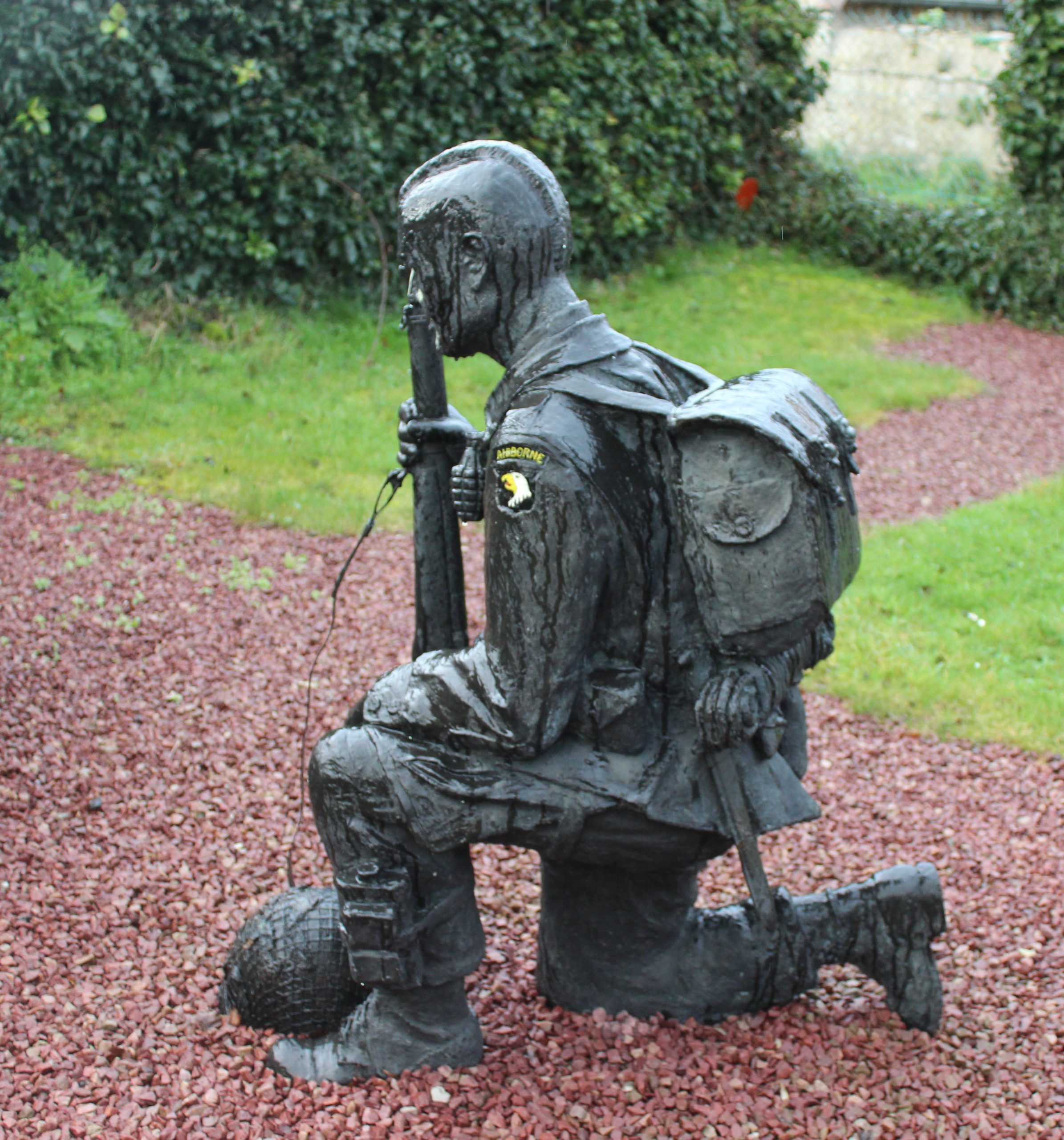A sculpture of a kneeling soldier in full gear holding a rifle, displayed on a bed of reddish-brown gravel with a green hedge in the background.