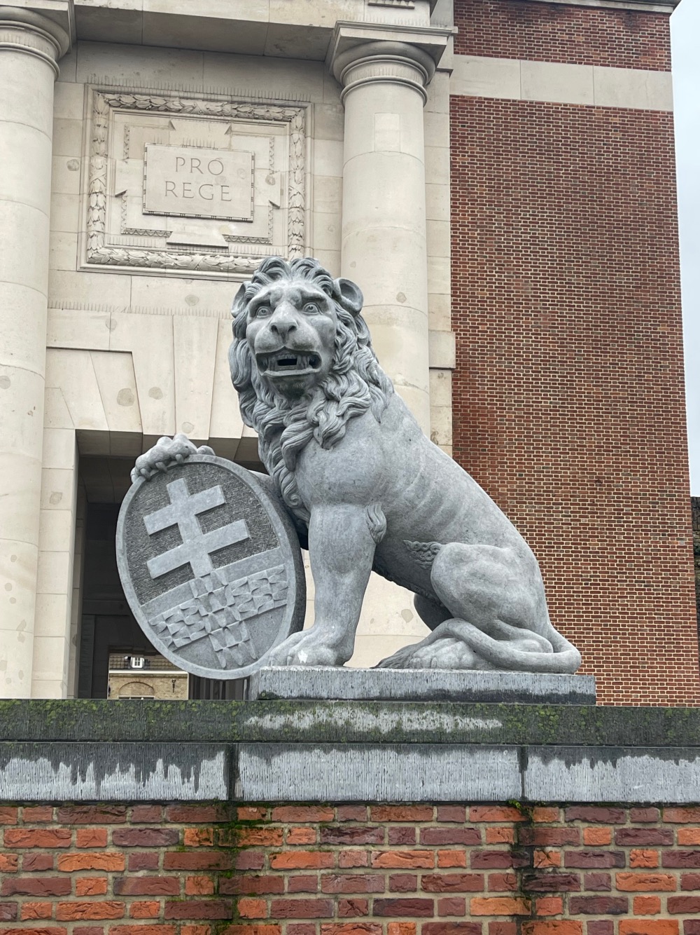 A stone sculpture of a lion holding a shield, positioned prominently in front of a building with brick columns and a stone facade engraved with "pro rege.