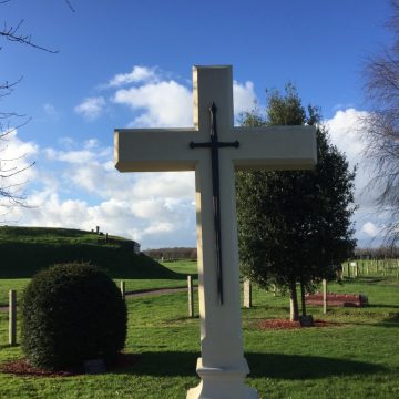 A large white cross with a smaller black cross on its surface, located in a lush green park with trees and a hill under a blue sky with scattered clouds.