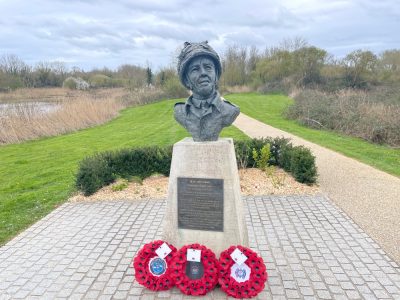 A bronze bust of a woman on a stone pedestal, adorned with remembrance poppy wreaths, overlooking a serene park with a walking path and a pond in the background.
