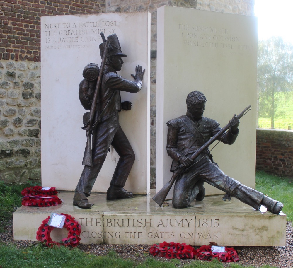 Sculpture depicting two british soldiers in dramatic poses from different eras, one standing with a rifle from 1815, the other kneeling with a modern rifle, at a memorial with inscriptions and wreaths.