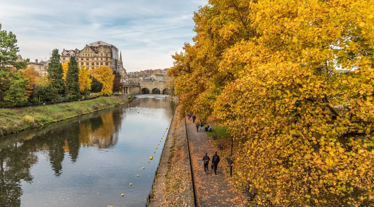 Autumn scene showing people walking along a tree-lined river path, with vibrant yellow foliage, overlooking a historic stone bridge and grand buildings in the background.
